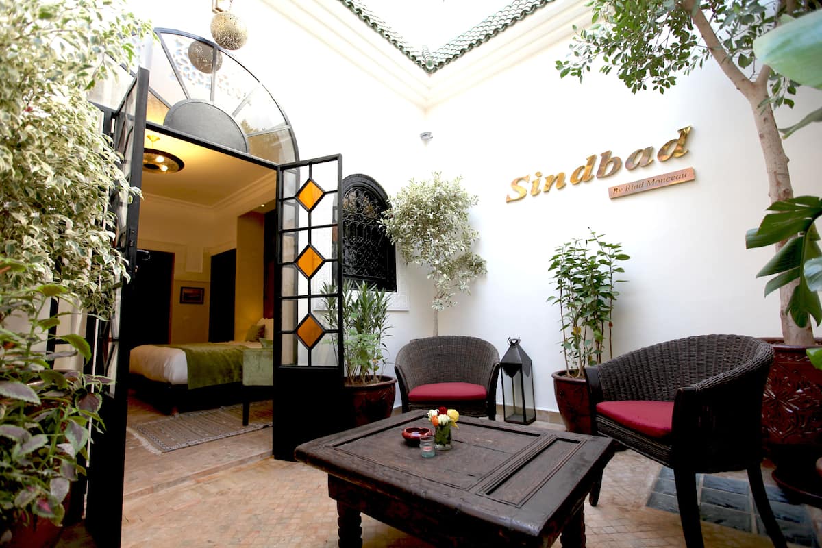 A room with a bed, a wicker chair, a table with a vase, and a wall with the name "Sindbad".