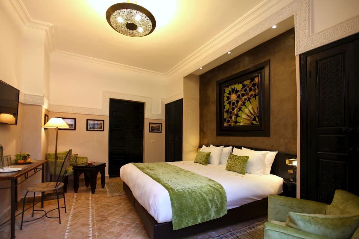 A hotel room with a bed, a desk, a chair, a lamp, and a painting on the wall.