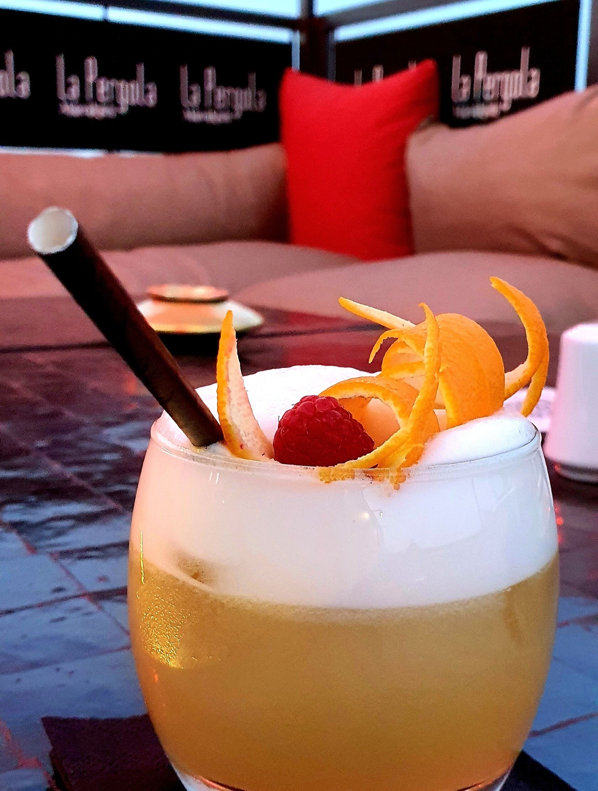 Ein Cocktail mit Fruchtverziierung auf einem Tisch in einem Raum, im Hintergrund sieht man eine Couch und Kissen.