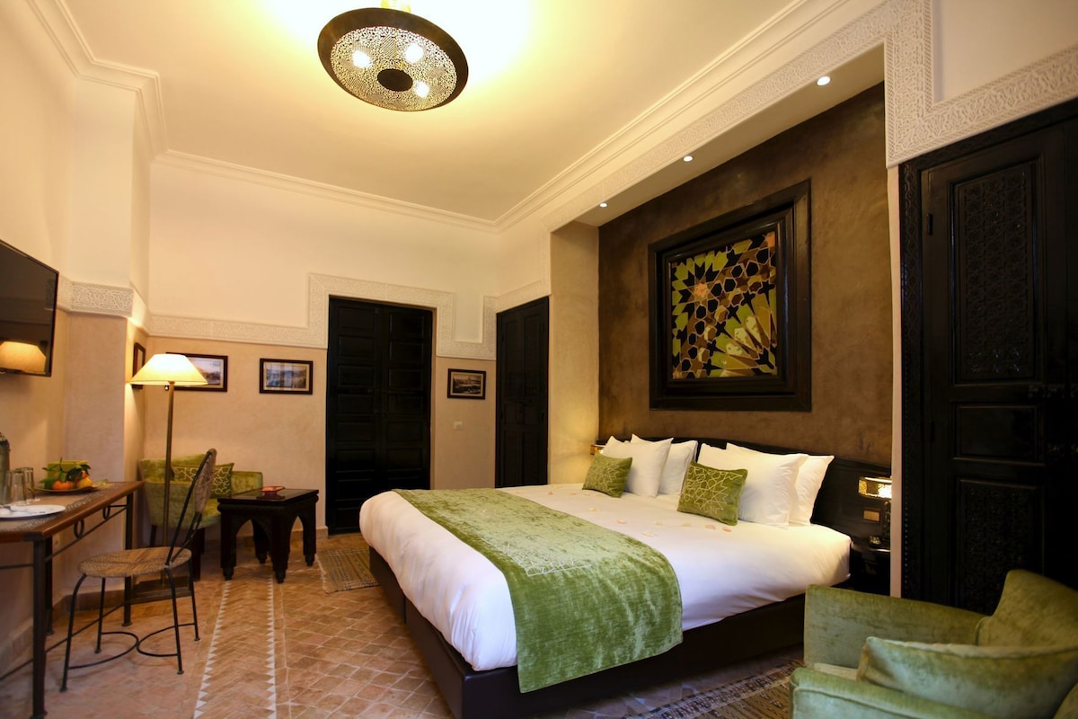 A hotel room with a bed, a desk, a chair, a lamp, and a painting on the wall.