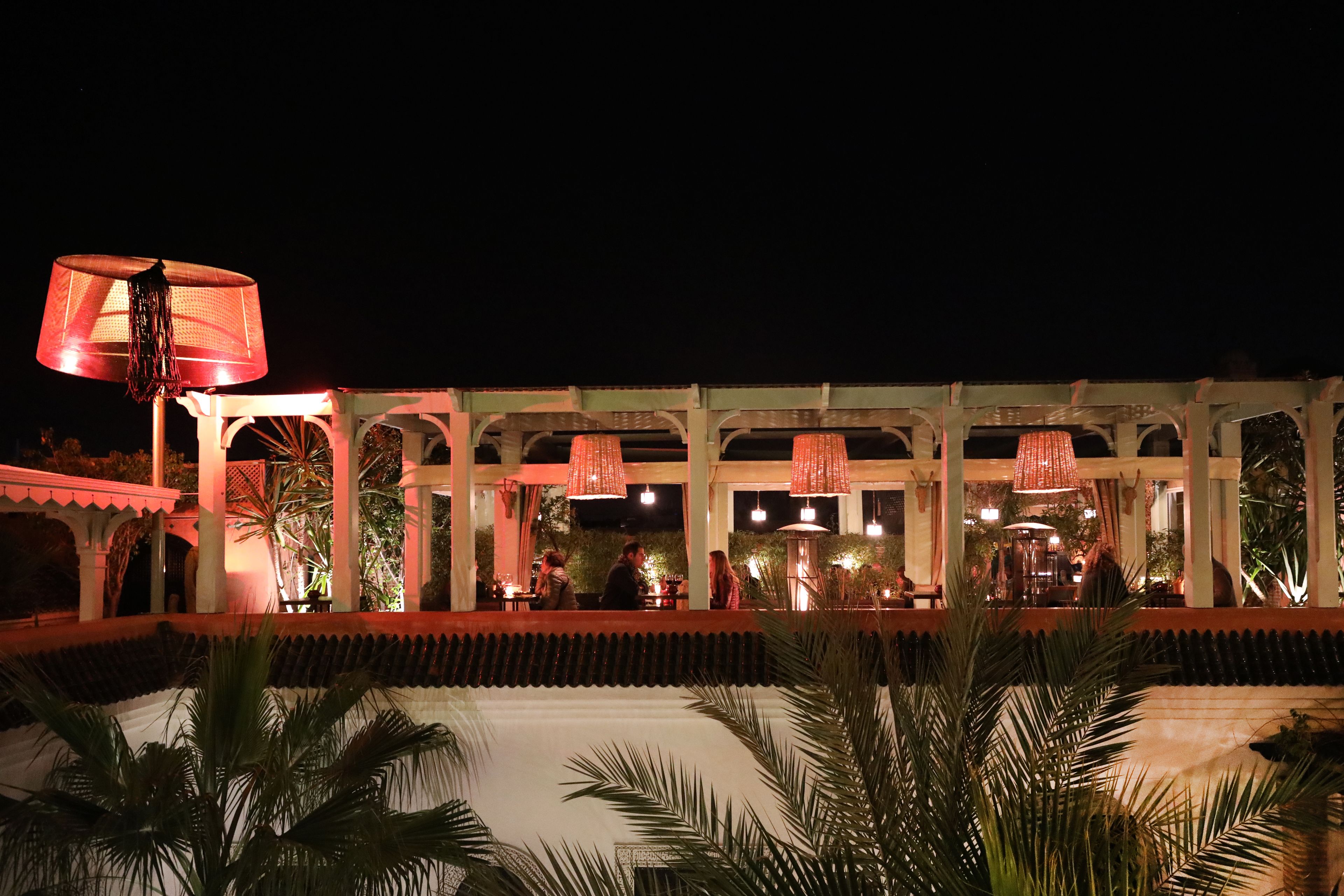 An outdoor dining area with a covered seating arrangement, palm trees, and hanging red lamps.