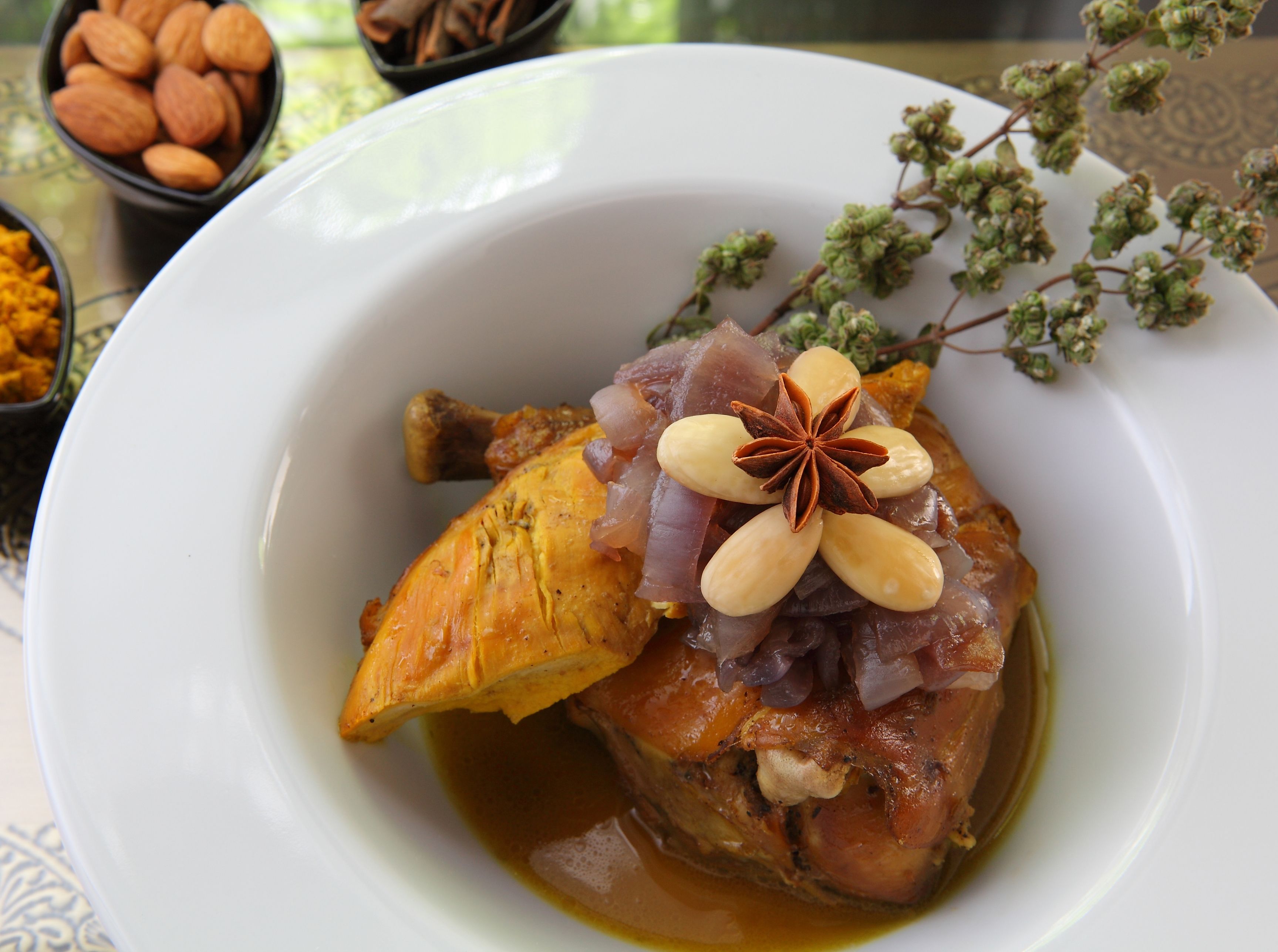 Un plato blanco con un muslo de pollo asado, decorado con almendras y anís estrellado, acompañado de un pequeño tazón de almendras y hierbas secas al fondo.