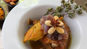 Un plato blanco con un muslo de pollo asado, decorado con almendras y anís estrellado, acompañado de un pequeño tazón de almendras y hierbas secas al fondo.