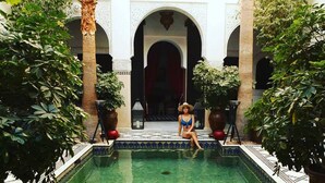 A pool area with a person sitting by the pool, surrounded by potted plants and architectural arches. - Le Riad Monceau (Marrakech)