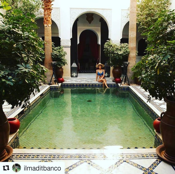 A pool area with a person sitting by the pool, surrounded by potted plants and architectural arches. - Le Riad Monceau (Marrakech)