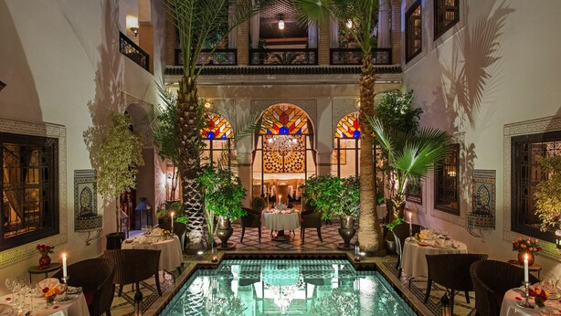 A luxurious indoor pool area with tables and chairs, surrounded by ornate architectural details and potted plants. - Le Riad Monceau (Marrakech)