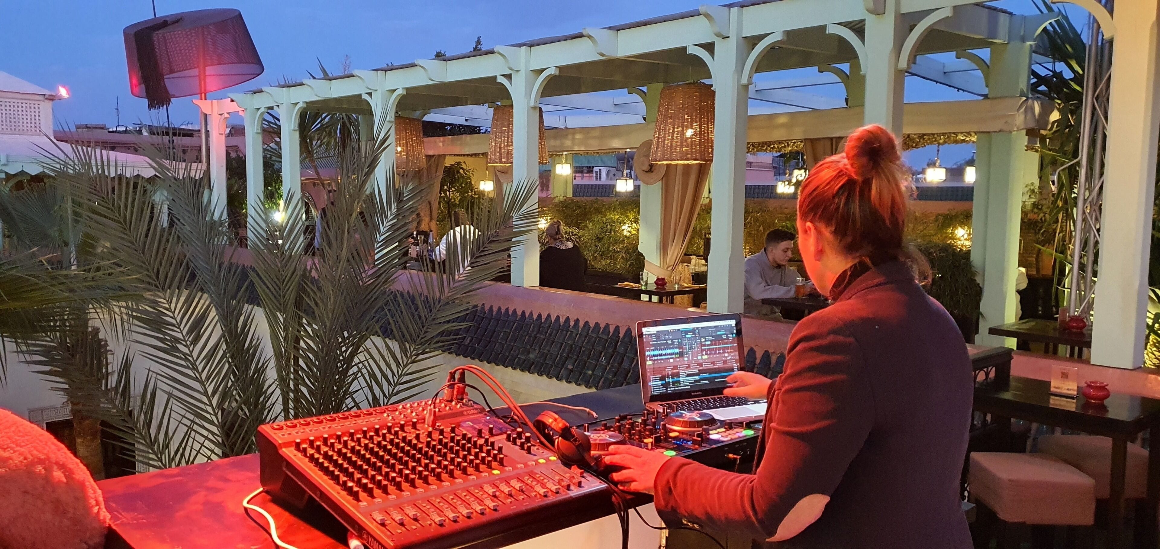 A person operating DJ equipment in an outdoor setting with a pergola, palm trees, and a view of buildings in the background.