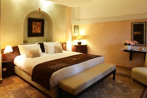 Una habitación de hotel con una cama grande, un banco, una mesita de noche con una lámpara, un espejo montado en la pared y un marco de fotos.