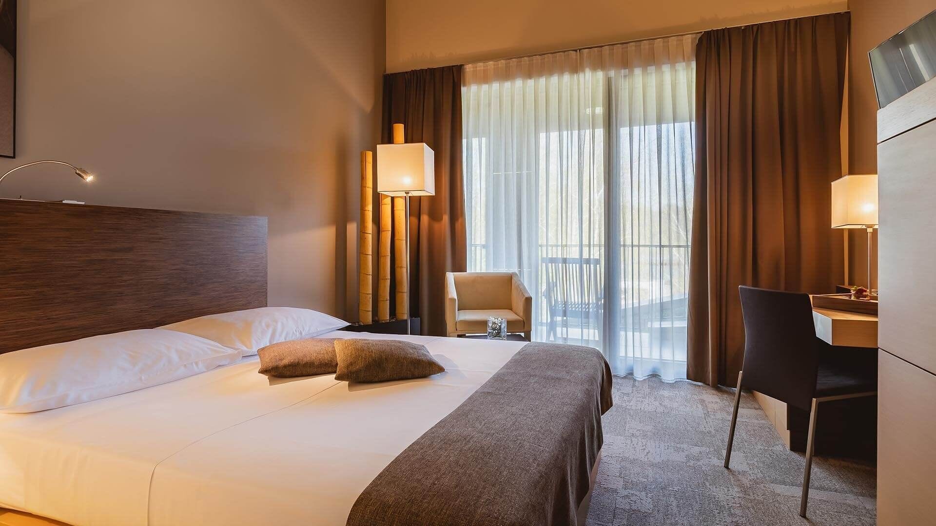 Een hotelkamer met een groot bed, twee bedlampen, een bureau met stoel, en uitzicht op het omliggende landschap door dunne gordijnen.