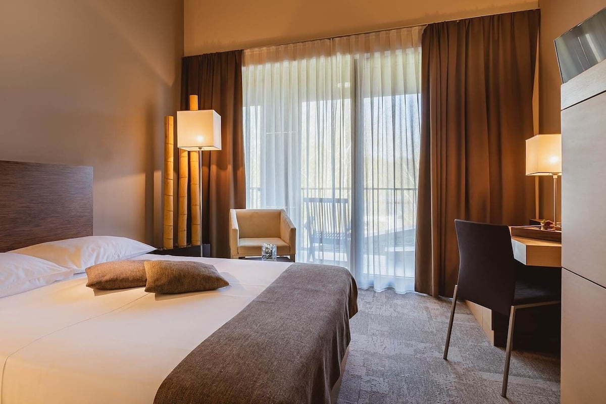Une chambre d’hôtel avec un grand lit, deux lampes de chevet, un bureau avec une chaise et une vue sur l’extérieur à travers des rideaux légers.