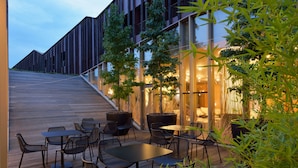 Un área de estar al aire libre con mesas y sillas, una terraza de madera y un edificio con ventanas amplias y una escalera.