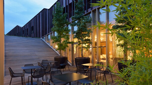 An outdoor seating area with tables and chairs, a wooden deck, and a building with large windows and a staircase. - Wellness Hotel Sotelia (Podcetrtek)