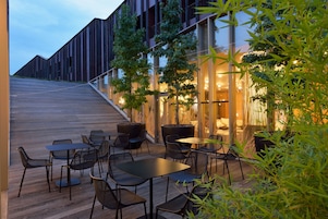 Un área de estar al aire libre con mesas y sillas, una terraza de madera y un edificio con ventanas amplias y una escalera.