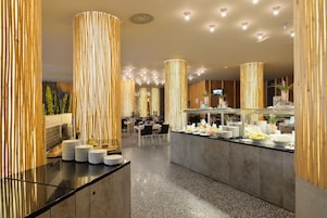 Un moderno vestíbulo de hotel con una recepción central, techos altos con iluminación empotrada y una zona de bufé con diversas opciones de comida.