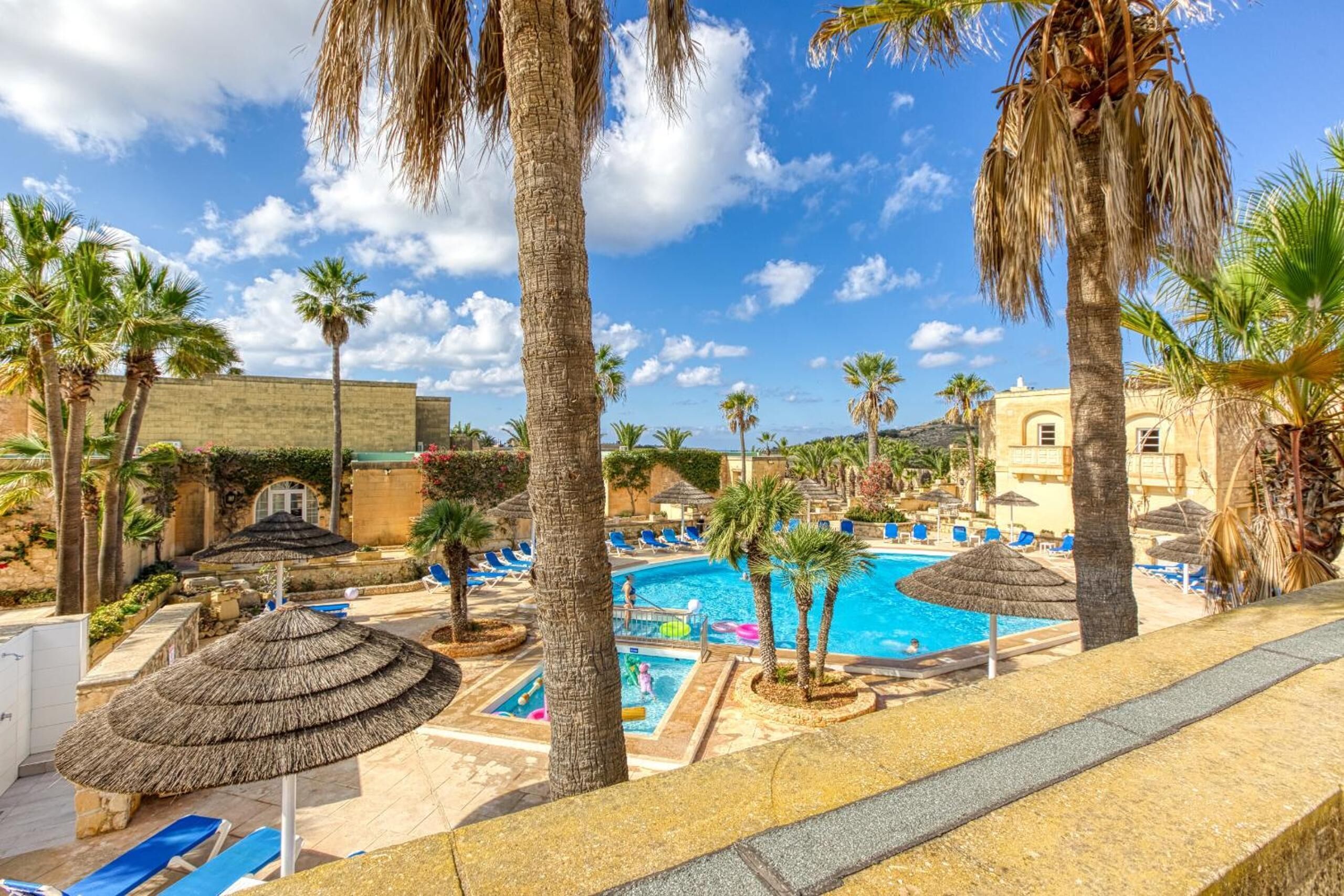 A pool area with lounge chairs, palm trees, and a building with a yellow facade.