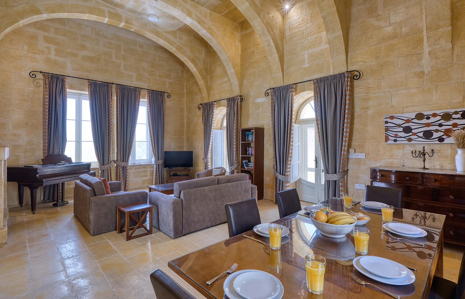 Un'ampia zona giorno con un pianoforte, divani, un tavolo da pranzo apparecchiato per la colazione e una parete in pietra con archi.