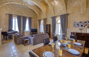 A spacious living area with a piano, sofas, a dining table set for breakfast, and a stone wall with arches. - Villagg Tal Fanal (Ghasri)