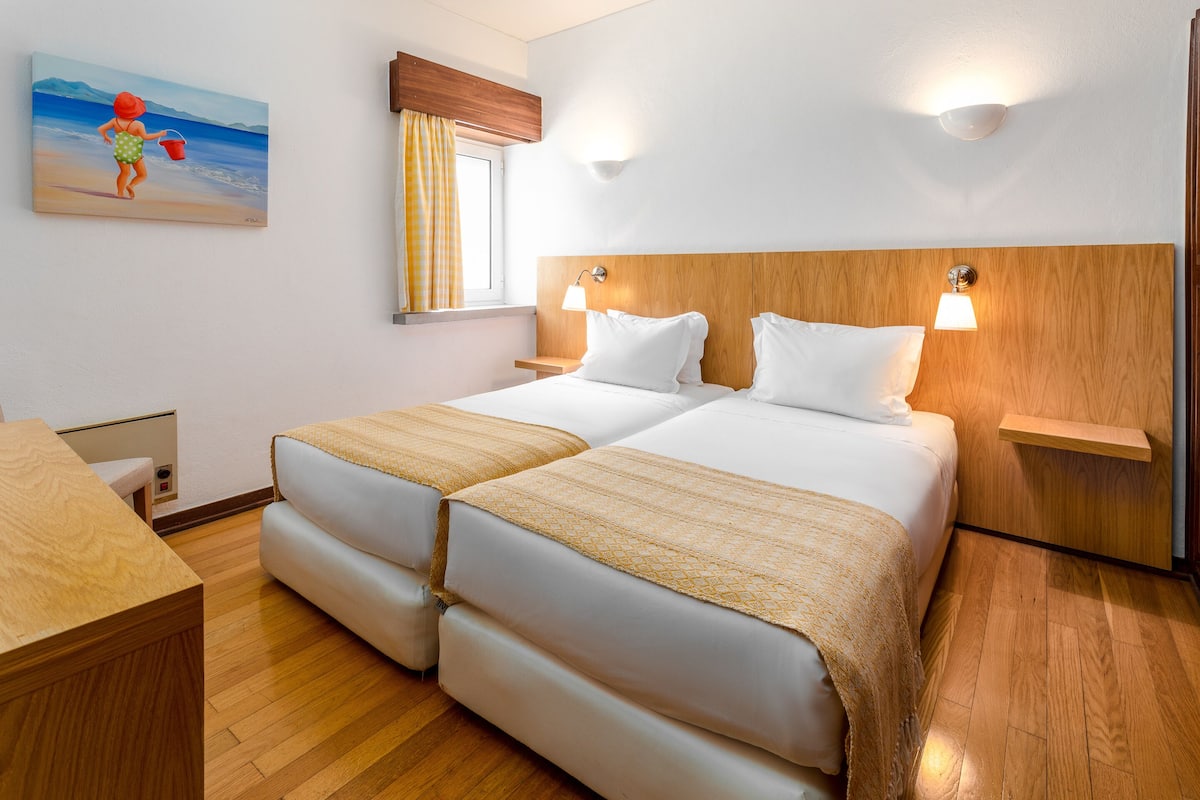A double bed with two pillows, a wooden headboard, bedside lamps, a wooden desk, and a painting of a beach scene.
