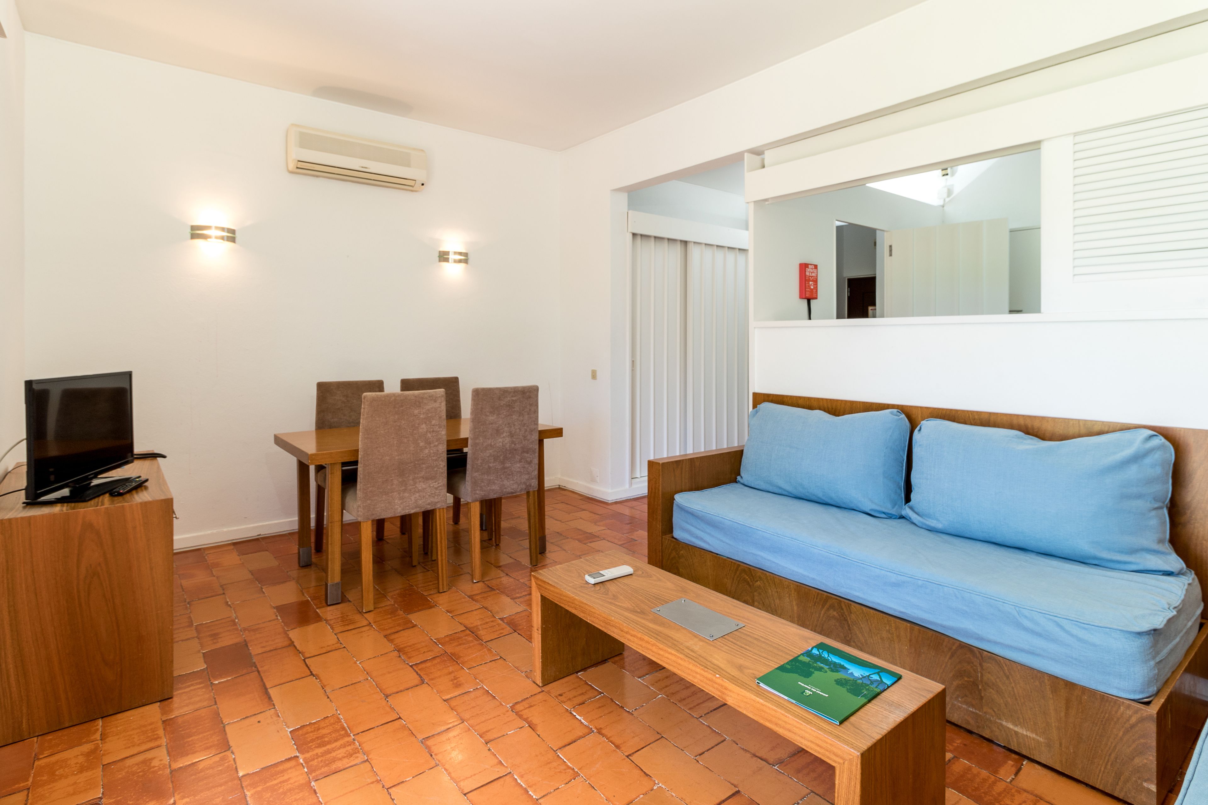 Uma sala de estar moderna com piso de madeira, um sofá azul, uma mesa de centro de madeira, uma TV em um móvel de madeira e uma área de refeições com cadeiras e uma mesa.
