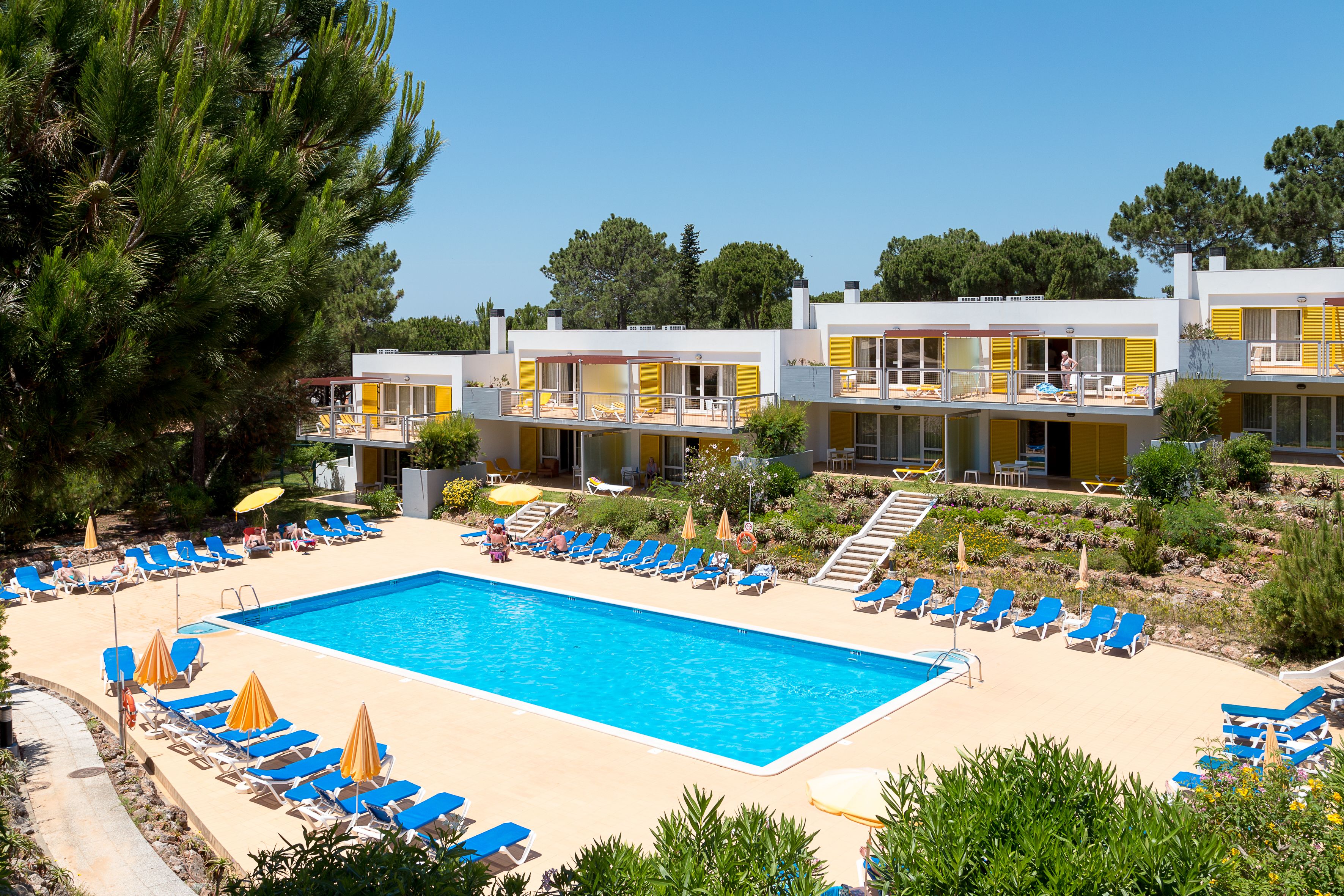 Ein Poolbereich mit blauen Liegestühlen und gelben Sonnenschirmen, umgeben von einem Gebäude mit Balkonen und blauem Himmel.