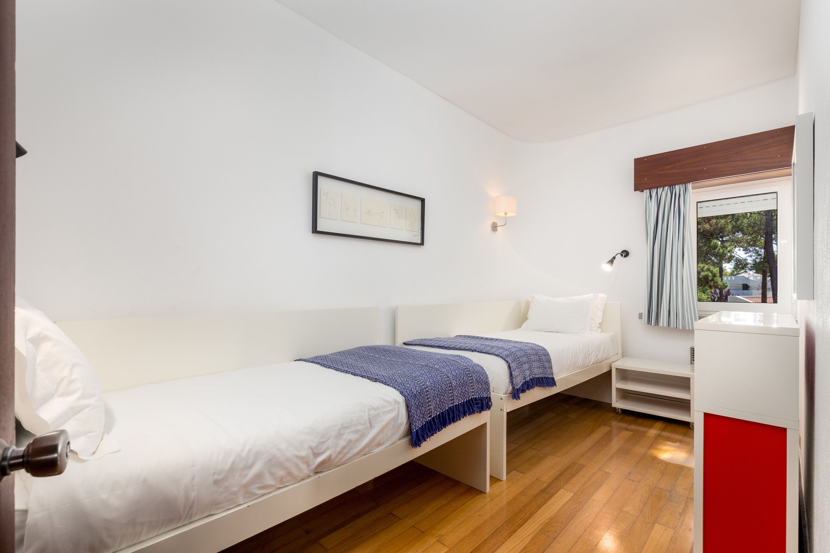 Une chambre moderne et minimaliste, dotée de deux lits simples, d’une œuvre d’art encadrée au mur, d’une table de chevet et d’une fenêtre avec des rideaux.
