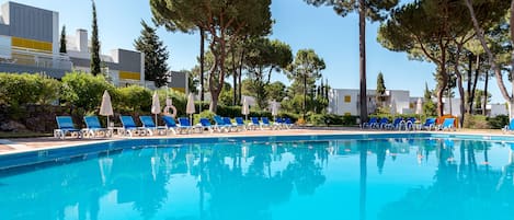 A swimming pool with blue lounge chairs and umbrellas, surrounded by trees and buildings.