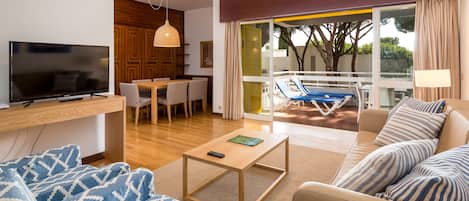 A modern living room with a flat-screen TV, a wooden coffee table, a sofa with patterned cushions, a dining area with chairs, and a balcony with a pool and lounge chairs.