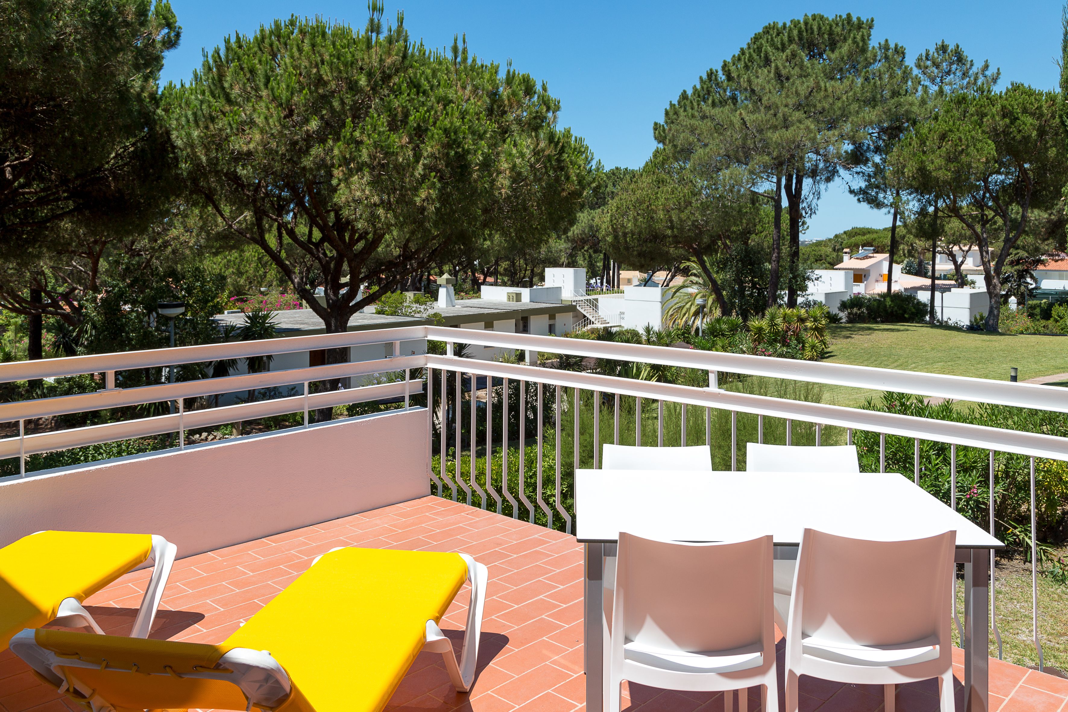 A terrace with yellow lounge chairs and a table with white chairs, overlooking a residential area with trees and houses.