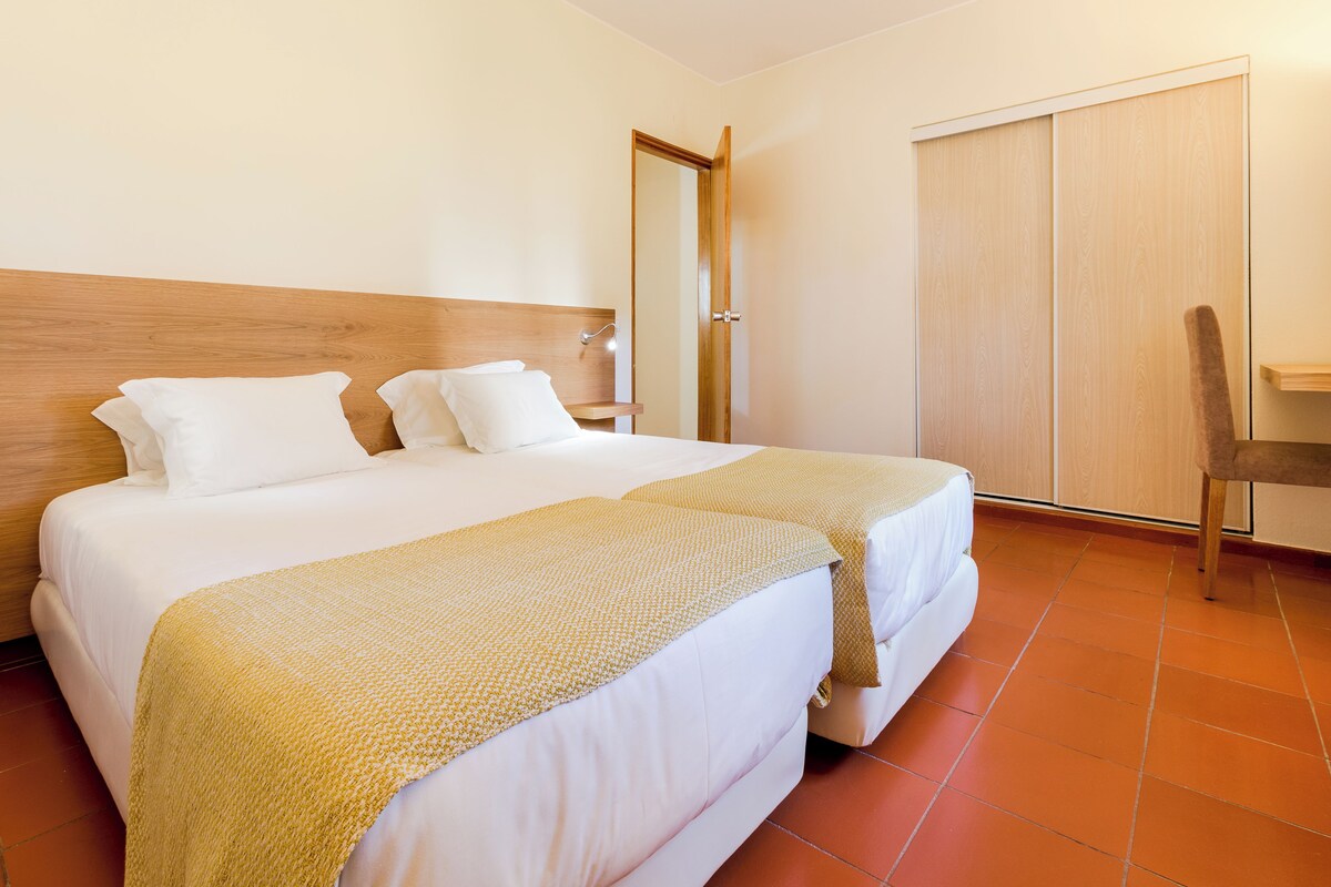 A hotel room with two beds, a wooden headboard, a bedside table with a lamp, a wardrobe, and a chair.