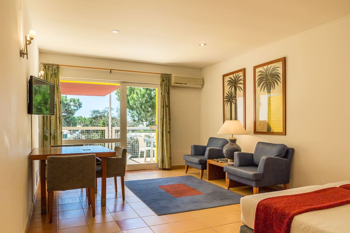 A hotel room with a balcony, a dining area, two armchairs, a sofa, a TV, and two framed pictures.