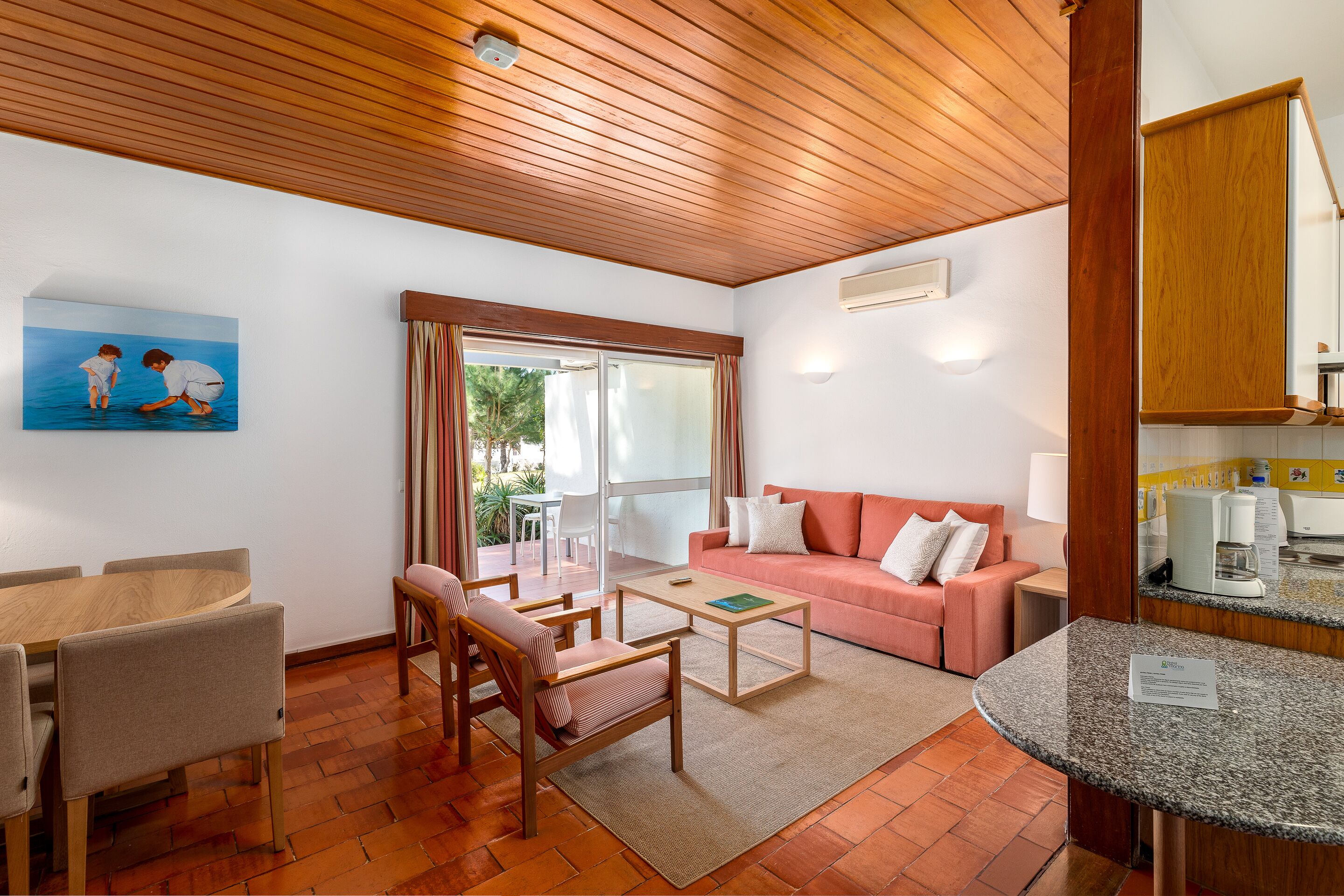 A cozy living room with a wooden ceiling, a pink sofa, a dining table with chairs, and a kitchen area with a counter and appliances.