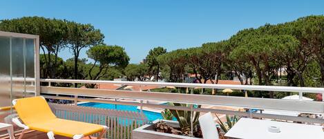 A poolside area with lounge chairs and a table, surrounded by trees and a clear sky.