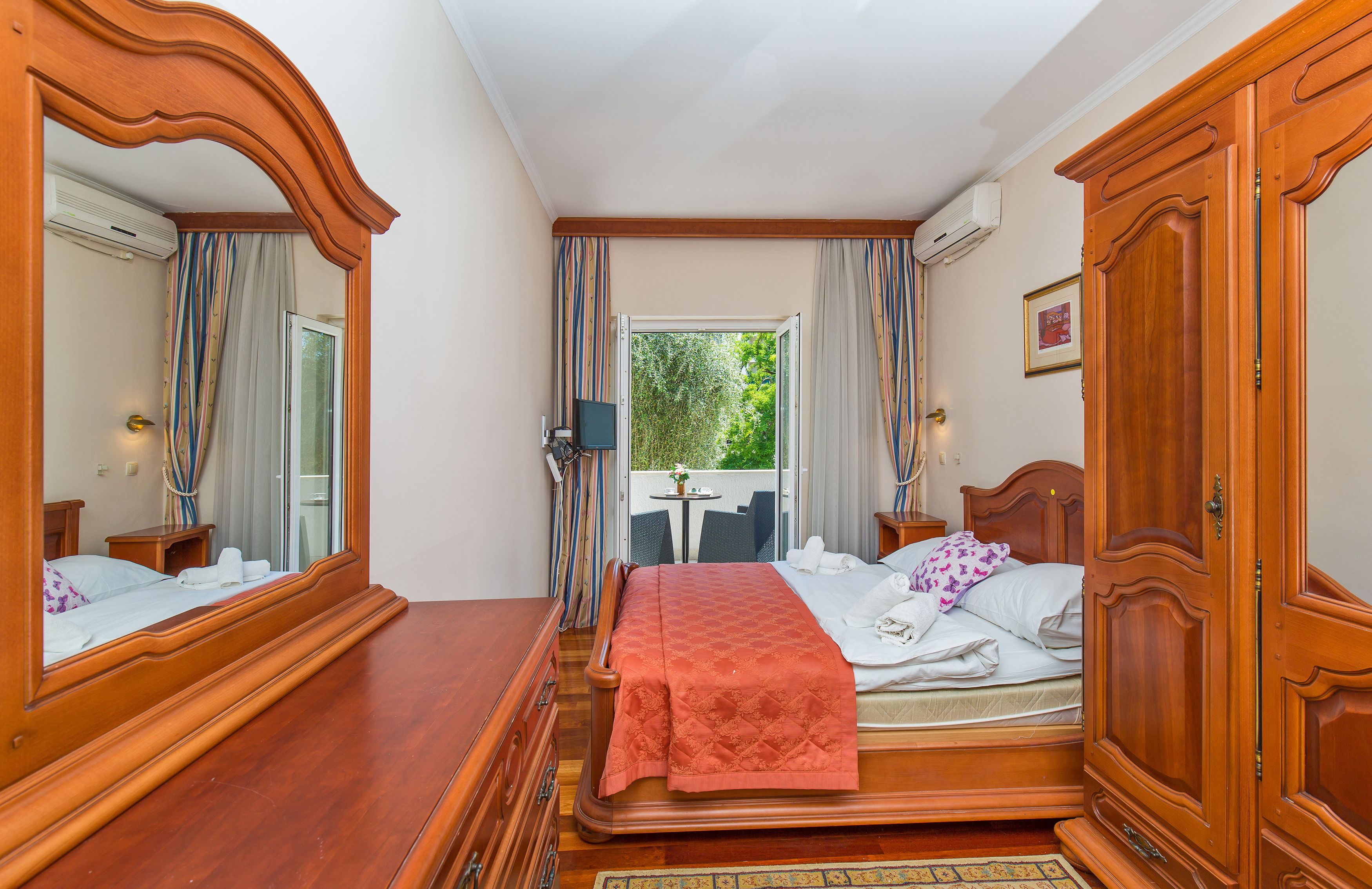 Ein Schlafzimmer mit einem hölzernen Bett, einem großen Spiegel, einem Kleiderschrank und Blick ins Freie durch ein Fenster.