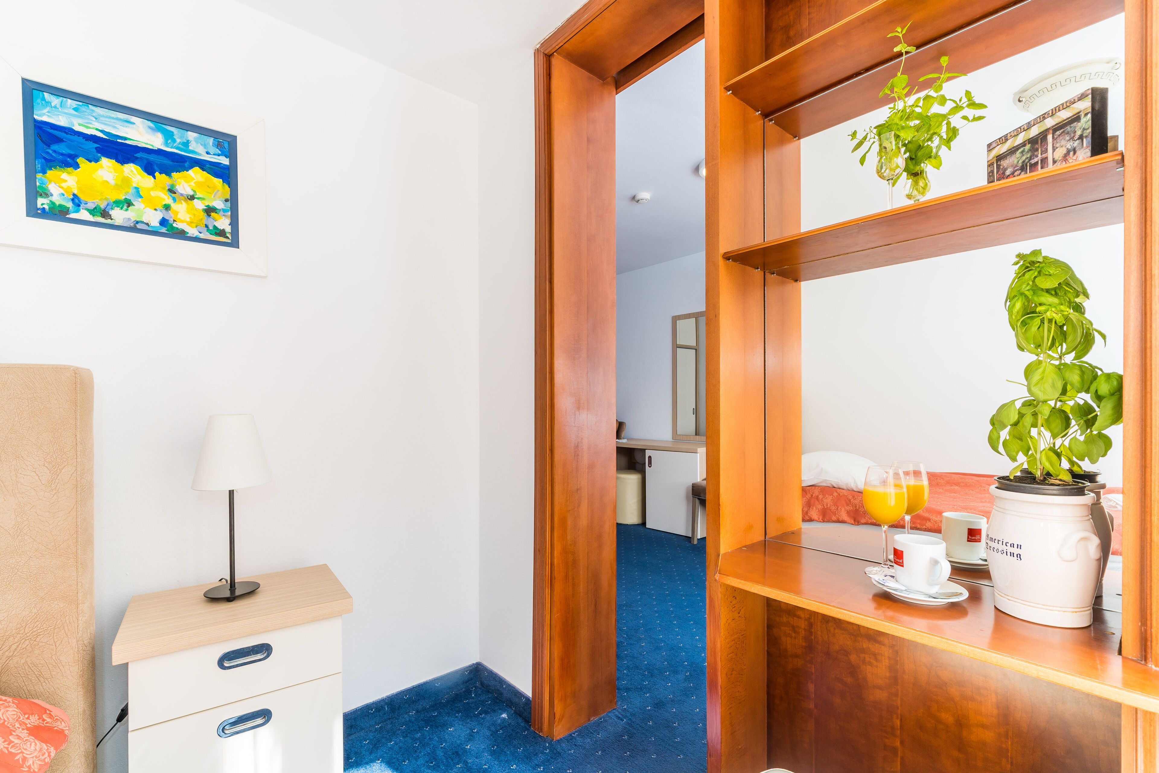 Habitación de hotel con un escritorio de madera, una mesita de noche con lámpara, una cama, una cafetera y una planta en maceta.
