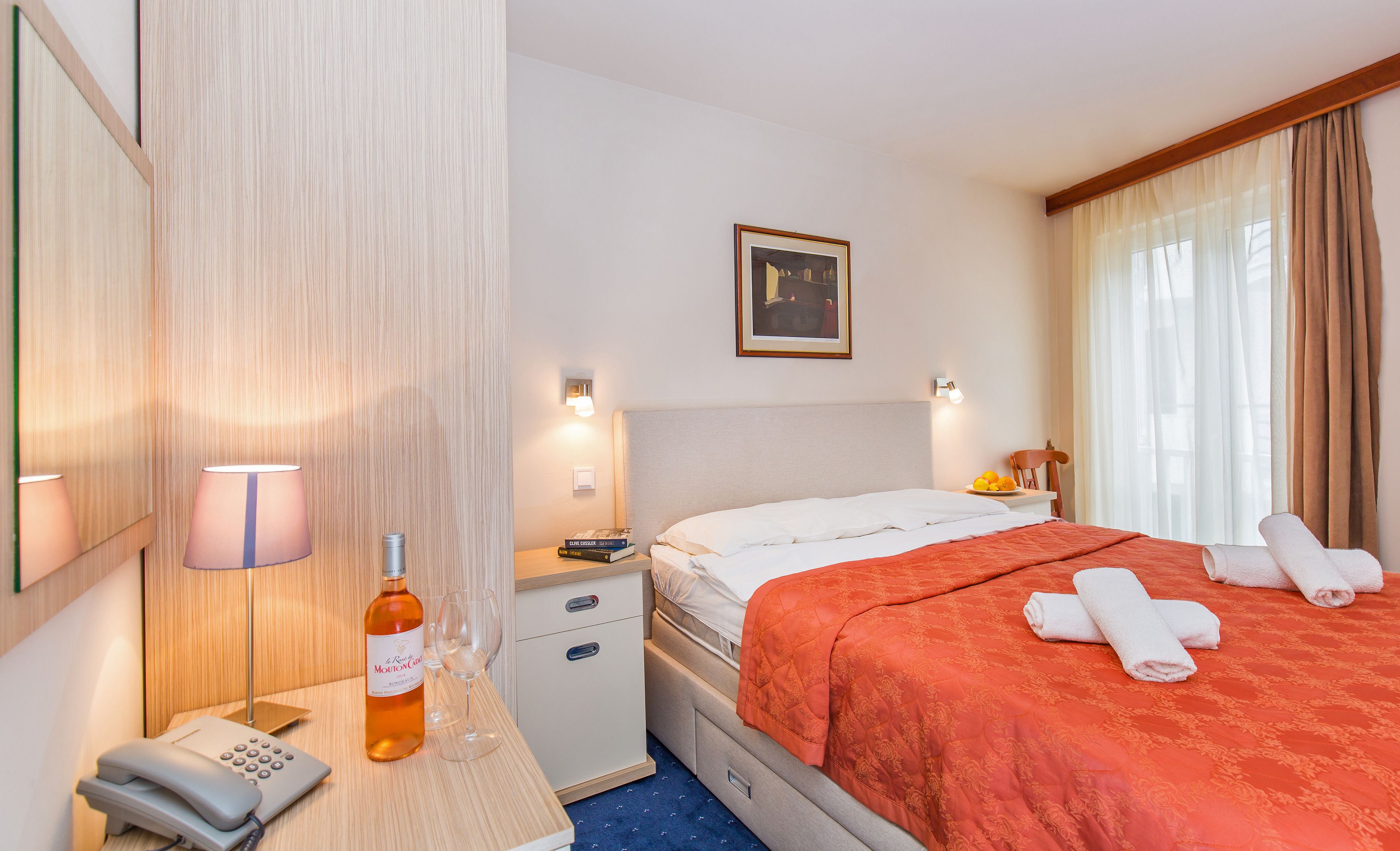 A hotel room with a bed, bedside table, lamp, phone, and a bottle of wine.