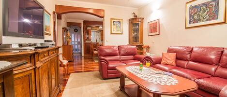 A spacious living room with a large flat-screen TV, leather sofas, a wooden coffee table, and a dining area in the background.