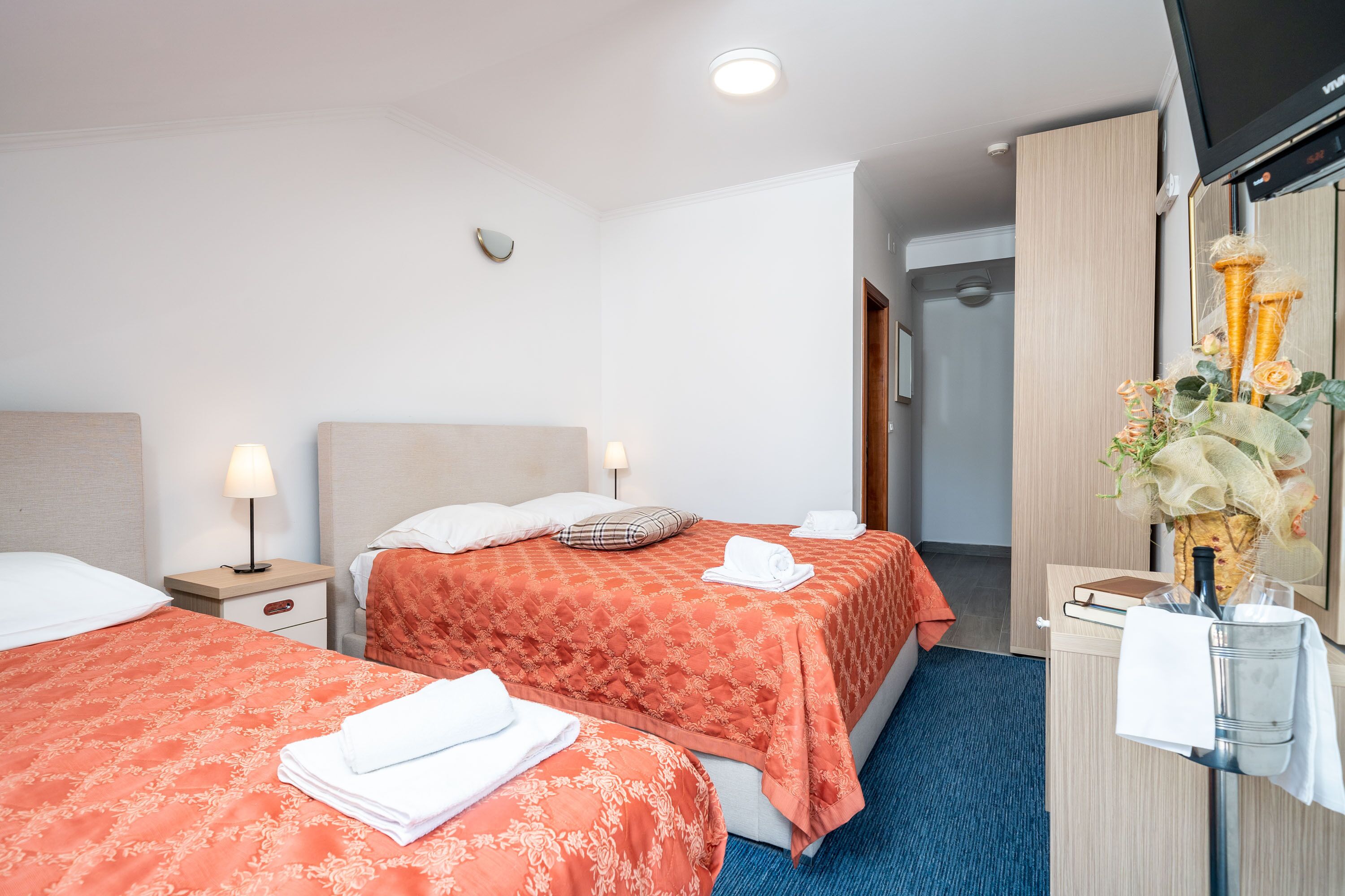 A hotel room with two beds, each with a red patterned bedspread and white pillows. There is a nightstand with a lamp, a small table with a vase of flowers, and a mirror on the wall.