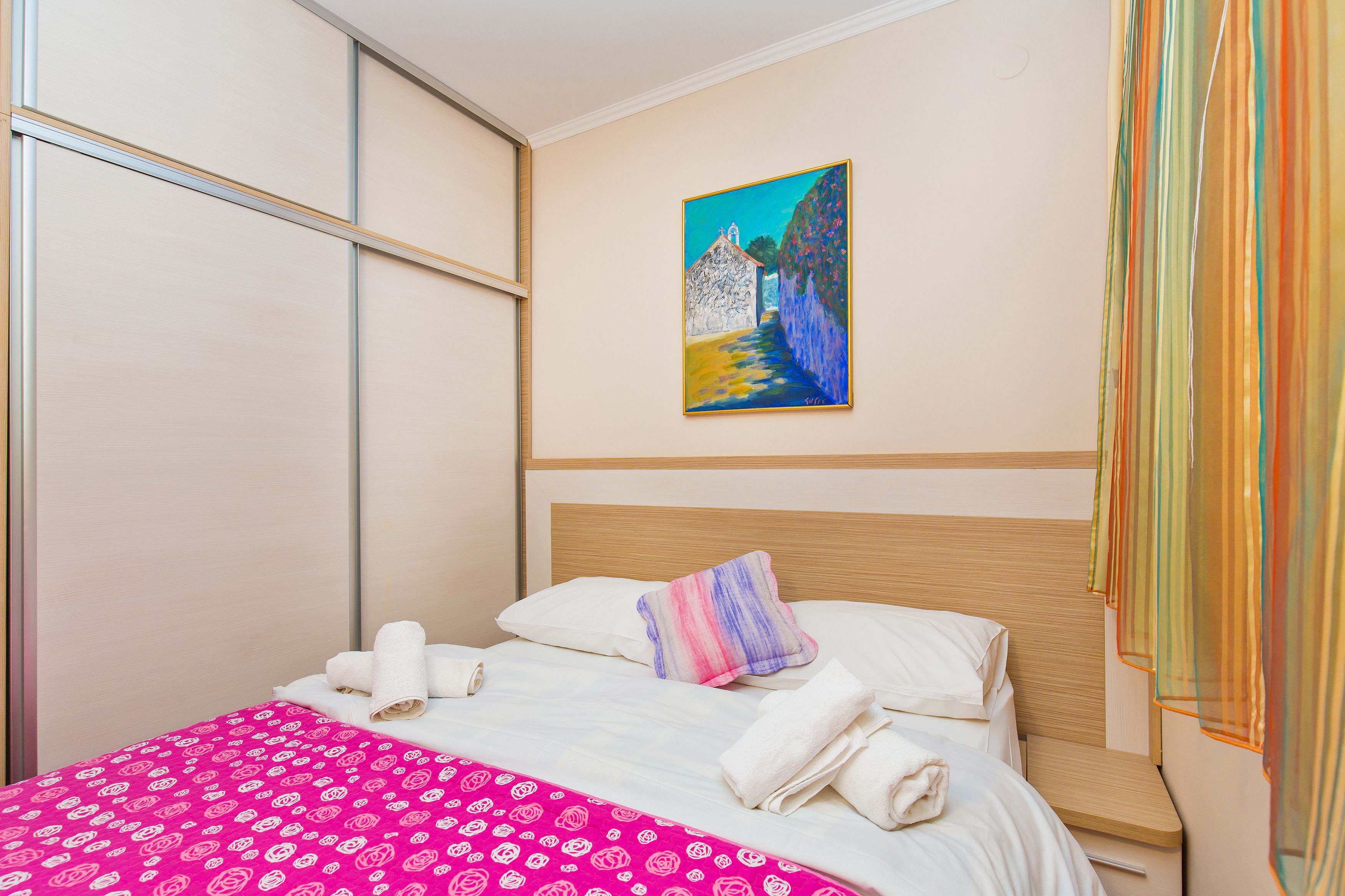 Een net opgemaakt bed met witte lakens en een roze geborduurde sprei, een schilderij van een landschap aan de muur, en een raam met dunne gordijnen.