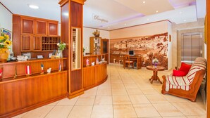 A hotel reception area with a wooden reception desk, a reception sign, a wall with a historical mural, a round table with a vase, and a sofa with red cushions. - Hotel Dubrovnik (Dubrovnik)