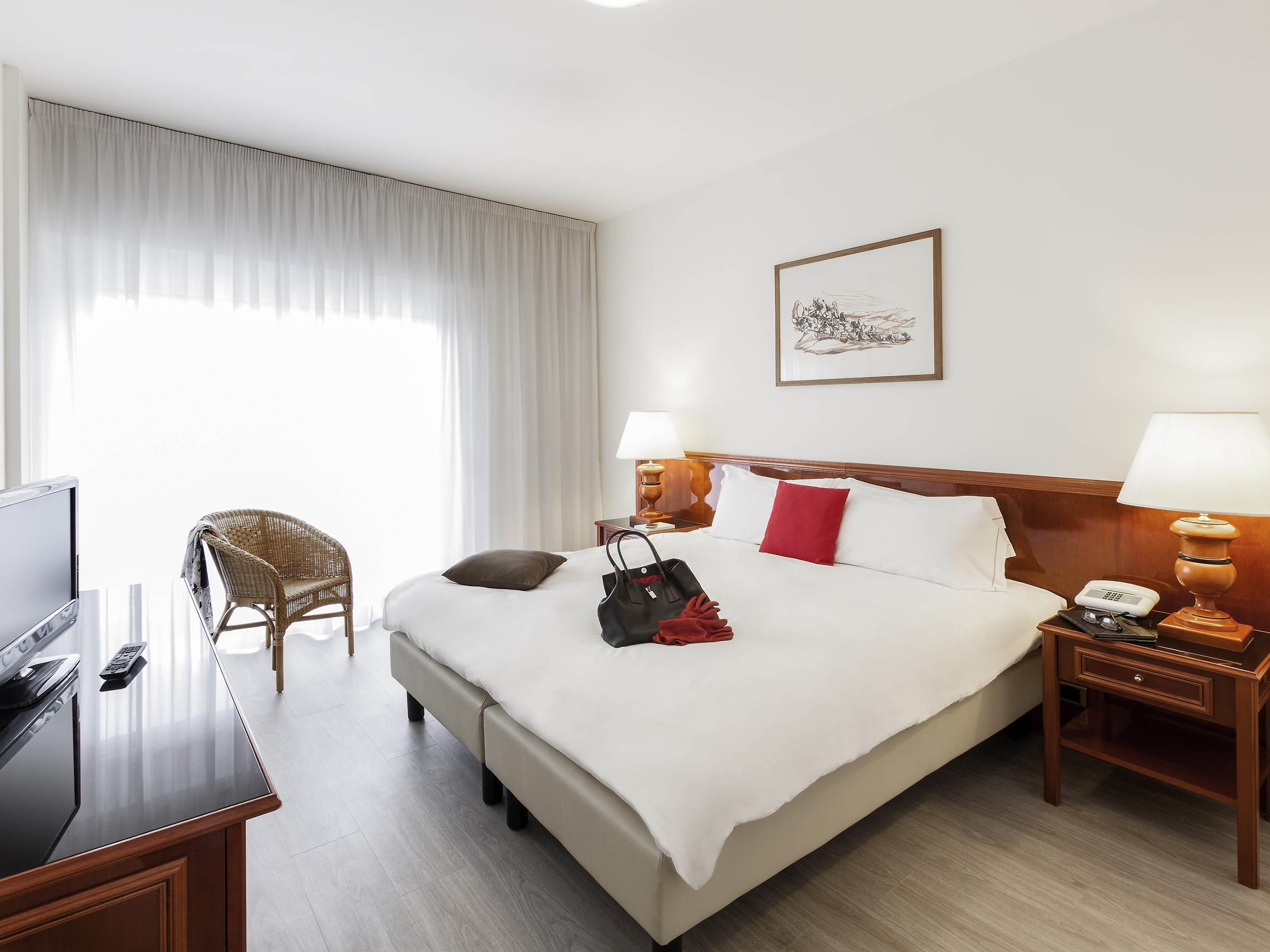 A hotel room with a large bed, two bedside lamps, a wooden nightstand, a wicker chair, a desk with a TV and remote, and a window with sheer curtains.