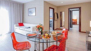 Una sala de estar moderna con una mesa de comedor de vidrio, sillas rojas, un sofá blanco con un cojín rojo y un ventanal con cortinas blancas.
