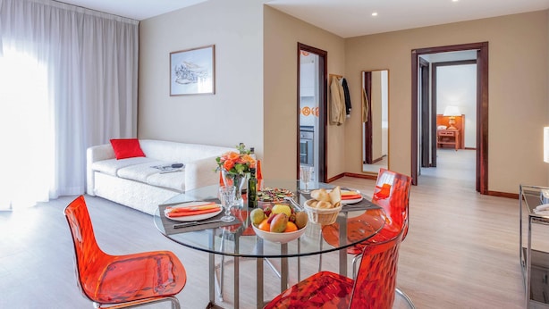 Un salon moderne avec une table à manger en verre, des chaises rouges, un canapé blanc avec un coussin rouge et une grande fenêtre ornée de rideaux blancs.