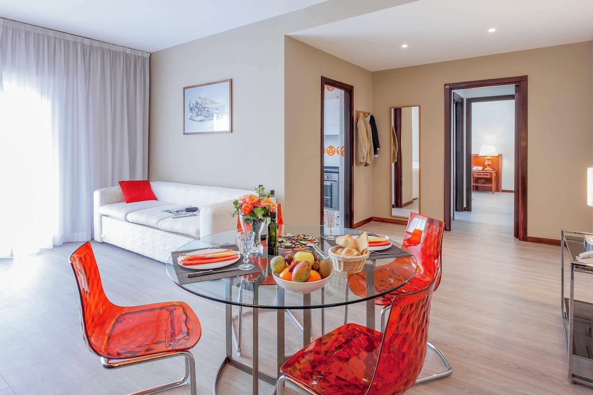 Un salon moderne avec une table à manger en verre, des chaises rouges, un canapé blanc avec un coussin rouge et une grande fenêtre ornée de rideaux blancs.