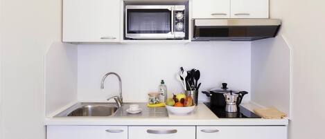 A compact kitchen with white cabinets, a stainless steel sink, a microwave, a stove, and a bowl of fruit on the countertop.