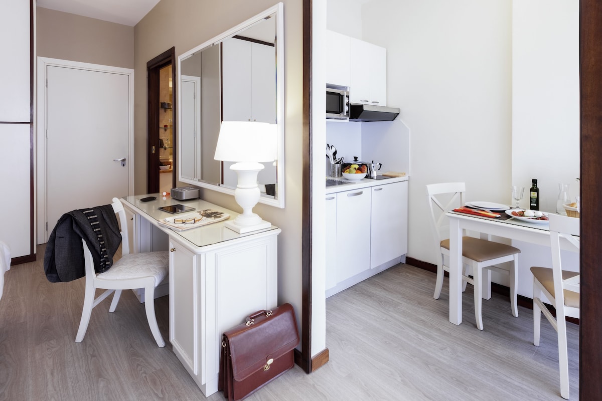 Une chambre d’hôtel compacte comprenant un coin cuisine, un bureau avec une chaise, une petite table à manger et un miroir.