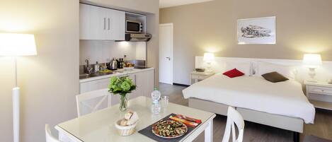 A hotel room with a bed, bedside tables, a small kitchenette, a dining table with chairs, and a vase with flowers.