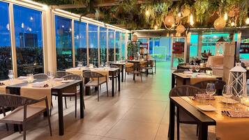 Restaurante com mesas montadas para refeições, grandes janelas e plantas penduradas.