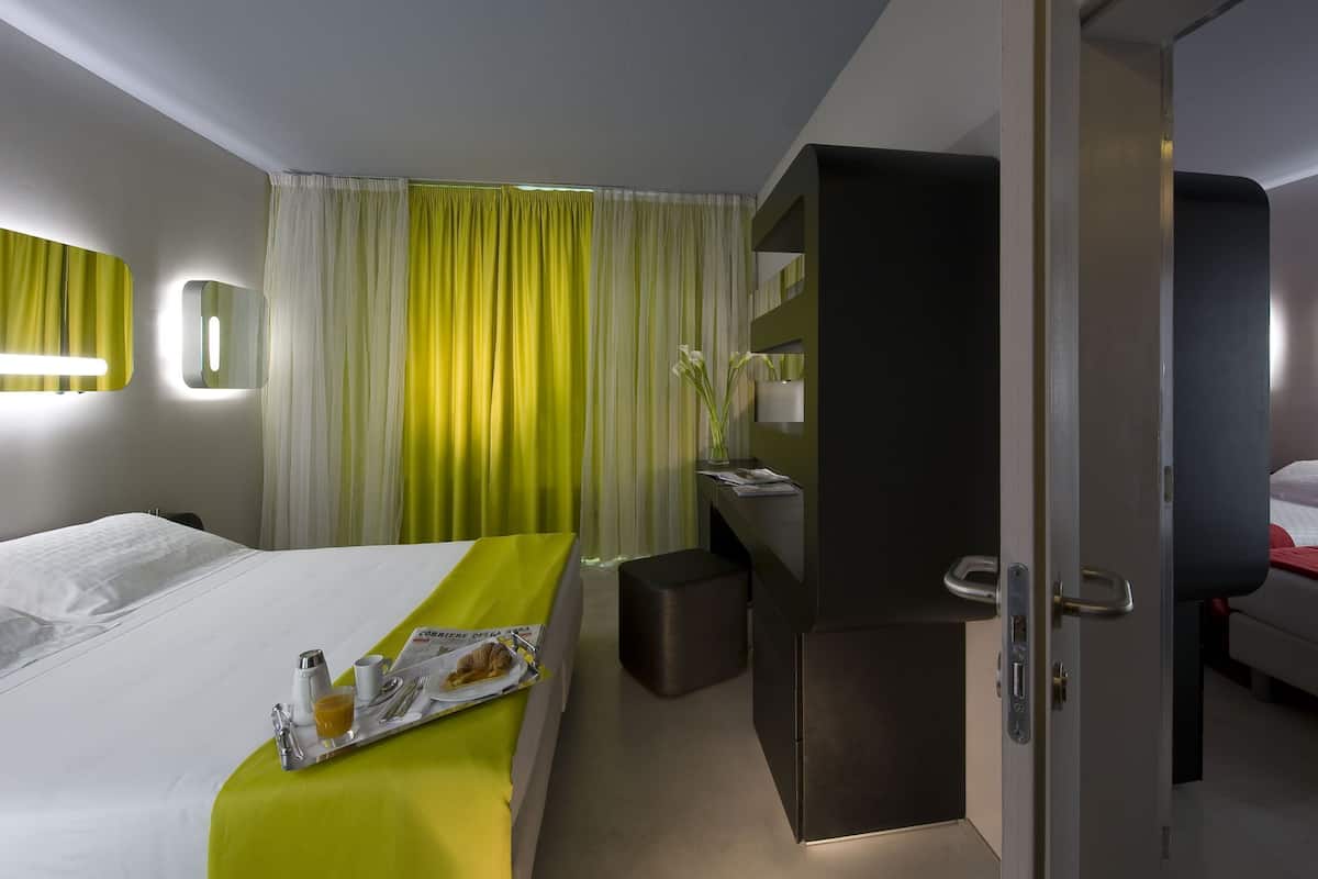 A hotel room with a bed, a desk, a chair, a small table with breakfast, and a door leading to another room.