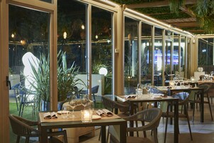 Un comedor interior con mesas dispuestas para dos, con sillas de mimbre y vistas al exterior a través de grandes ventanales.