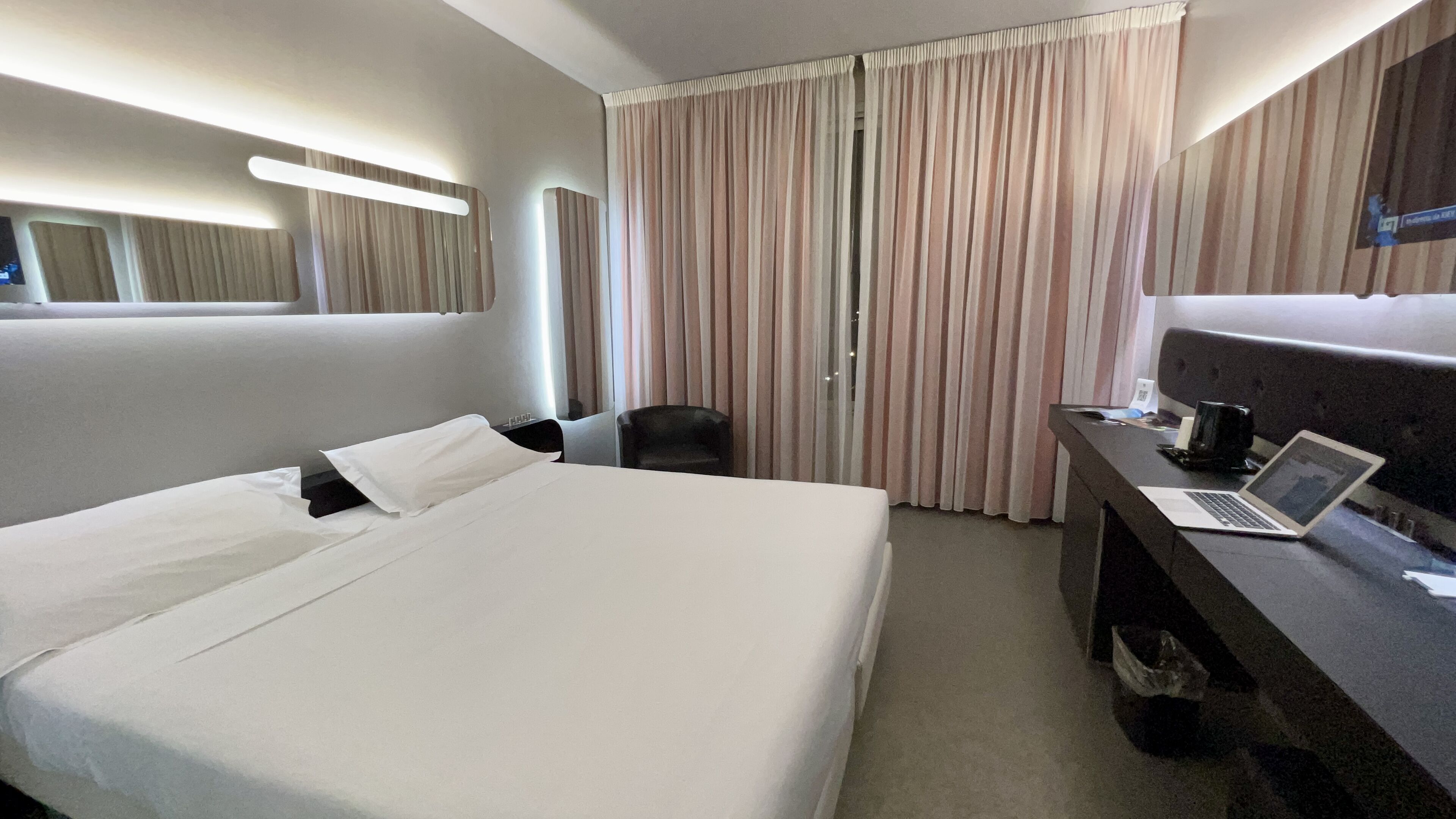A hotel room with a large bed, a desk with a laptop, a chair, and a window with curtains.
